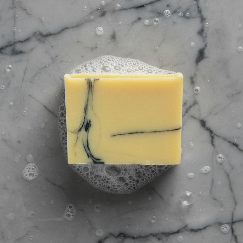 A bar of soap with a light color and black swirl patterns, placed on a gray and black marble surface that is wet with some bubbes