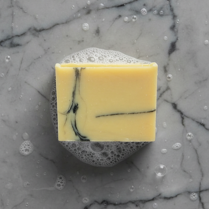 A bar of soap with a light color and black swirl patterns, placed on a gray and black marble surface that is wet with some bubbes
